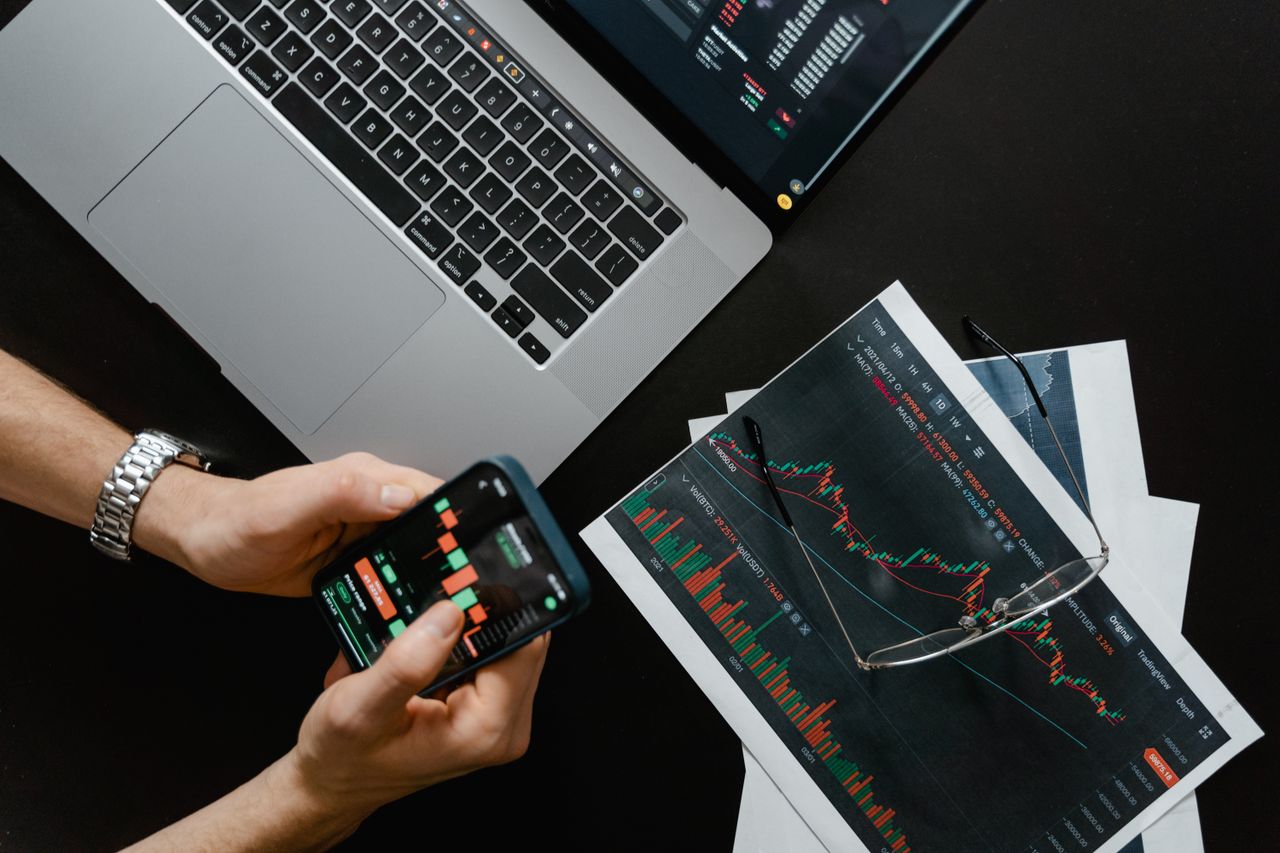 Des mains utilisant un smartphone à côté d’un ordinateur portable avec des graphiques boursiers, illustrant le trading numérique.